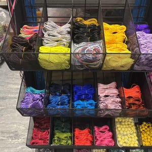 May include: A metal rack filled with colourful spools of thread, labelled "Rattaail". The spools are organised by colour, with some colours having multiple spools. There are also bags of beads in various colours.