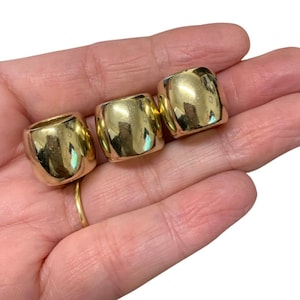 May include: Three gold-coloured, square-shaped beads with rounded edges. The beads are smooth and reflective, with a slight indentation on each side. The beads are held in a hand.
