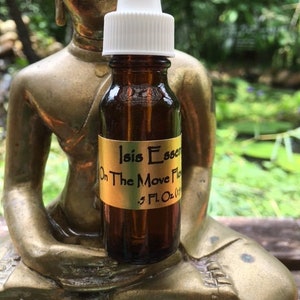 Puede incluir: Una pequeña botella de vidrio marrón con un cuentagotas blanco. La botella tiene una etiqueta dorada con el texto "Isis Essence On The Move Flow 5 Fl Oz (1)". La botella está colocada frente a una estatua dorada de Buda.