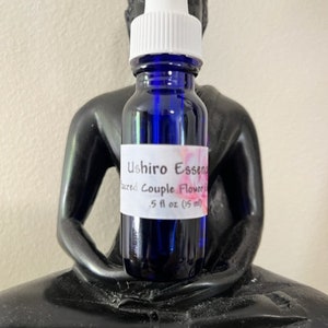 May include: A dark blue glass bottle with a white dropper top. The label reads "Ushiro Essence Sacred Couple Flower" and "5 fl oz (15 ml)". The bottle is set against a black statue of a seated Buddha.