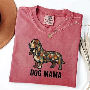 May include: A faded rose-coloured t-shirt with a basset hound illustration in a patchwork design. The text "DOG MAMA" is printed below the dog. A gold necklace with a pendant is draped across the shirt.