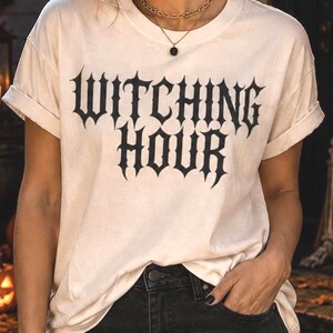 May include: A cream-colored short-sleeve t-shirt featuring the text "WITCHING HOUR" printed in a black, stylized, gothic-inspired font across the chest. The shirt has a crew neckline and rolled sleeves, paired with black denim jeans.