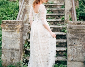 May include: A woman in a white lace wedding dress with a lace bodice and a long skirt. She is standing on a set of stone steps with greenery in the background.