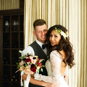 May include: A bride and groom pose for a wedding portrait. The bride is wearing a white lace dress with a low back and a floral crown. The groom is wearing a gray suit with a white shirt and a black bow tie. They are holding a bouquet of red, pink, and white flowers.