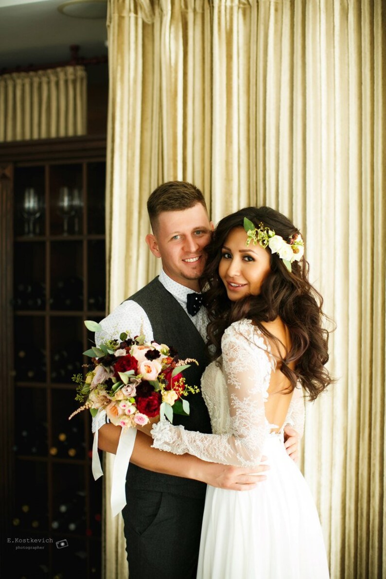 May include: A bride and groom pose for a wedding portrait. The bride is wearing a white lace dress with a low back and a floral crown. The groom is wearing a gray suit with a white shirt and a black bow tie. They are holding a bouquet of red, pink, and white flowers.