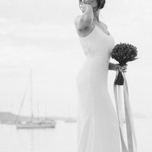 May include: A bride in a white sleeveless dress holds a bouquet of small white flowers with long pink and white ribbons.
