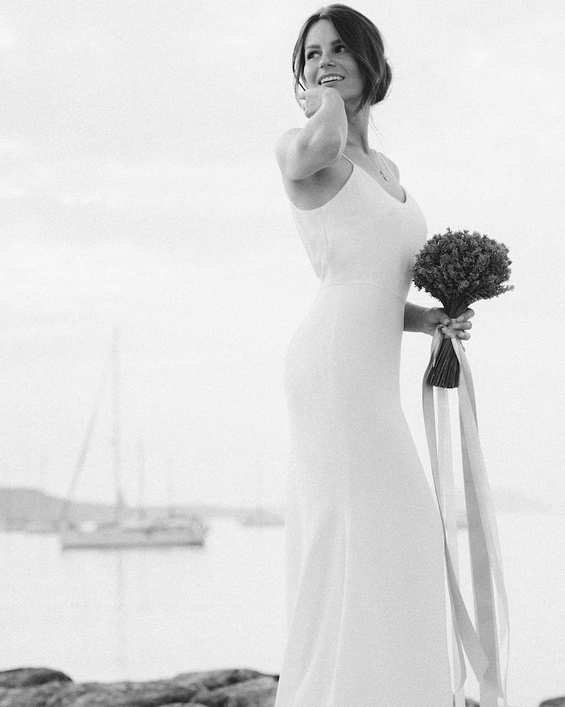 May include: A bride in a white sleeveless dress holds a bouquet of small white flowers with long pink and white ribbons.
