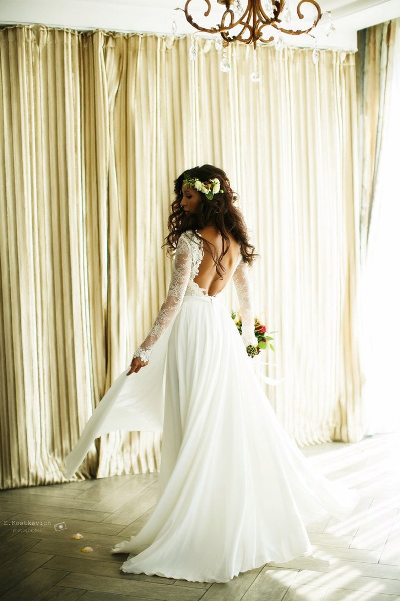 May include: A bride wearing a white lace wedding dress with a low back and long sleeves. She is wearing a flower crown and holding a bouquet of flowers. The dress has a flowing skirt and is photographed in a room with white curtains.