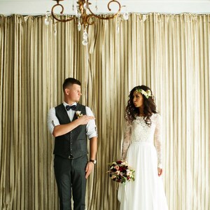 May include: A bride and groom stand in front of a gold curtain. The bride is wearing a white lace dress with a floral crown. The groom is wearing a gray suit with a vest and a bow tie. They are both smiling.