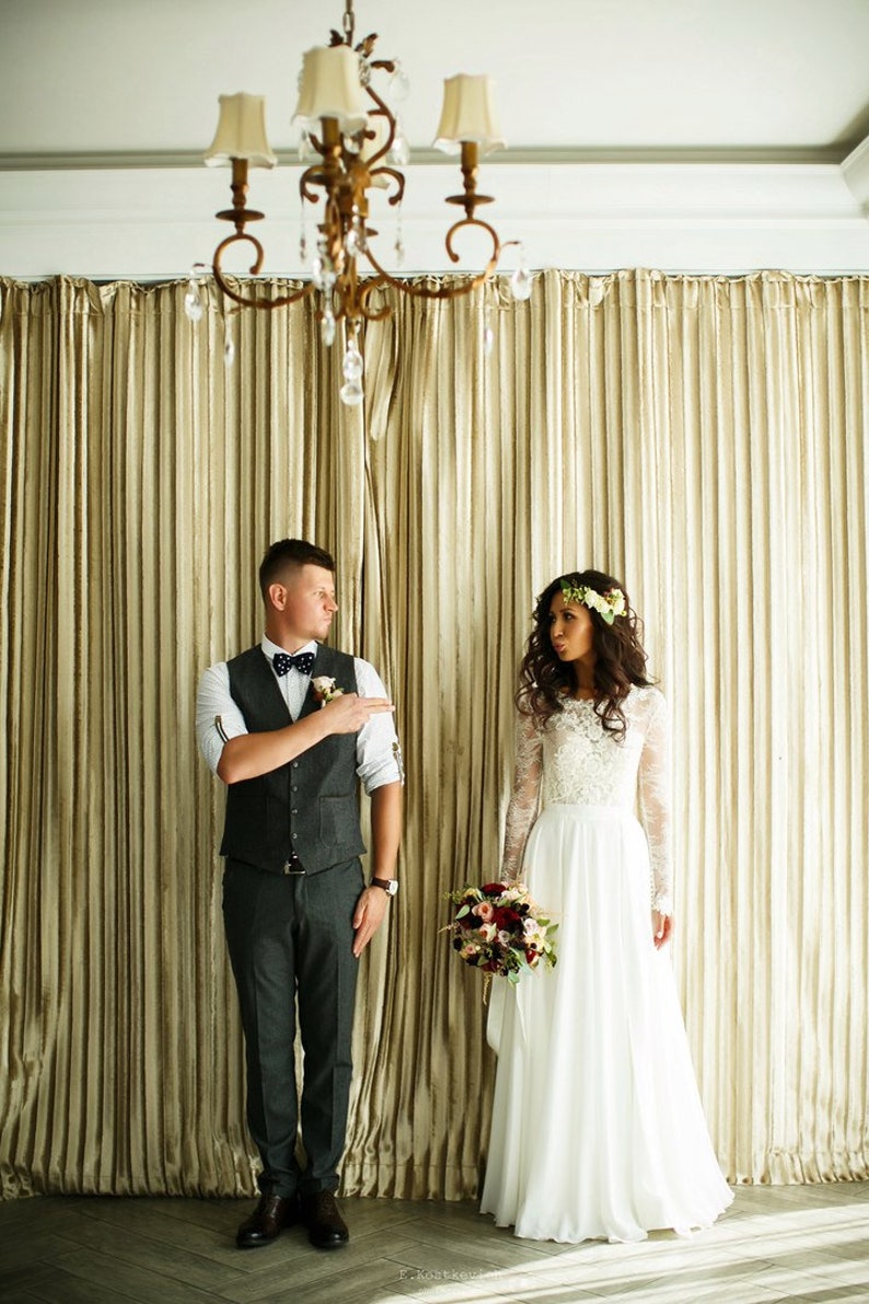 May include: A bride and groom stand in front of a gold curtain. The bride is wearing a white lace dress with a floral crown. The groom is wearing a gray suit with a vest and a bow tie. They are both smiling.