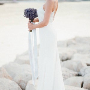May include: A bride in a white sleeveless gown holds a bouquet of lavender flowers. The bride is standing on a rocky beach.