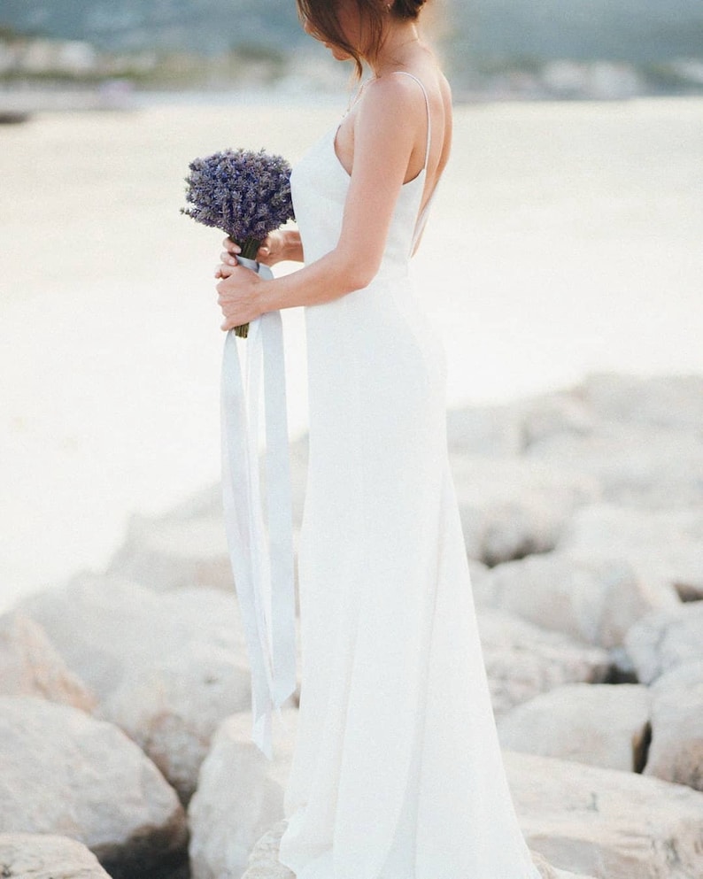 May include: A bride in a white sleeveless gown holds a bouquet of lavender flowers. The bride is standing on a rocky beach.