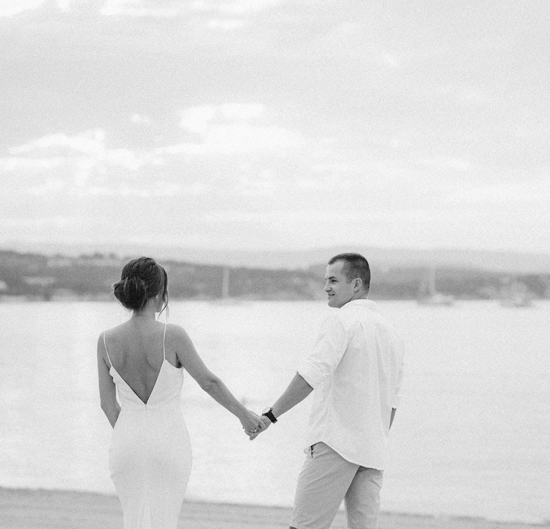 May include: A black and white photo of a couple holding hands and walking away from the camera on a beach.