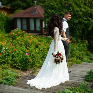 May include: A bride and groom walking down a wooden path in a garden setting. The bride is wearing a white wedding dress and carrying a bouquet of red and white flowers. The groom is wearing a gray suit and a white shirt. The path is surrounded by green bushes and flowers.