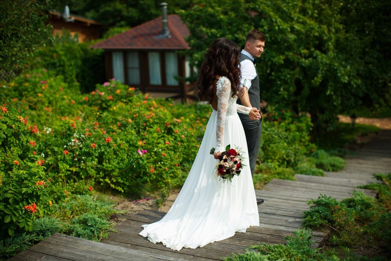 May include: A bride and groom walking down a wooden path in a garden setting. The bride is wearing a white wedding dress and carrying a bouquet of red and white flowers. The groom is wearing a gray suit and a white shirt. The path is surrounded by green bushes and flowers.