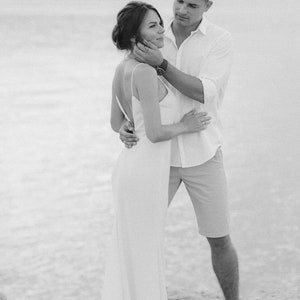 May include: A black and white photo of a couple embracing on a beach. The woman is wearing a white dress and the man is wearing a white shirt and shorts.