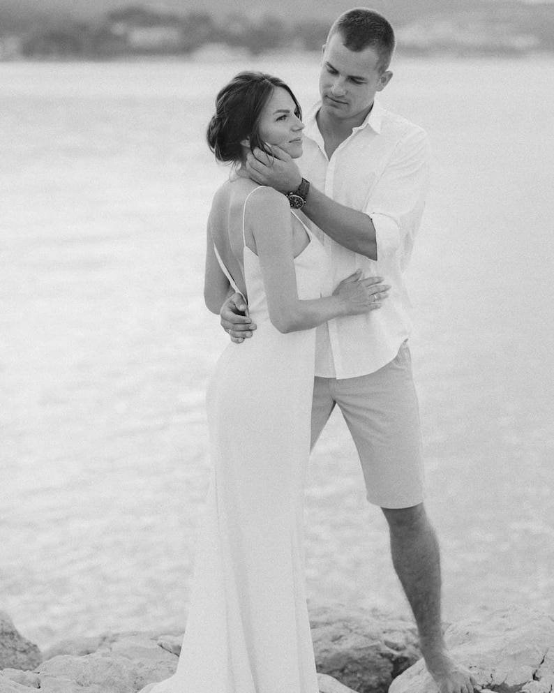 May include: A black and white photo of a couple embracing on a beach. The woman is wearing a white dress and the man is wearing a white shirt and shorts.
