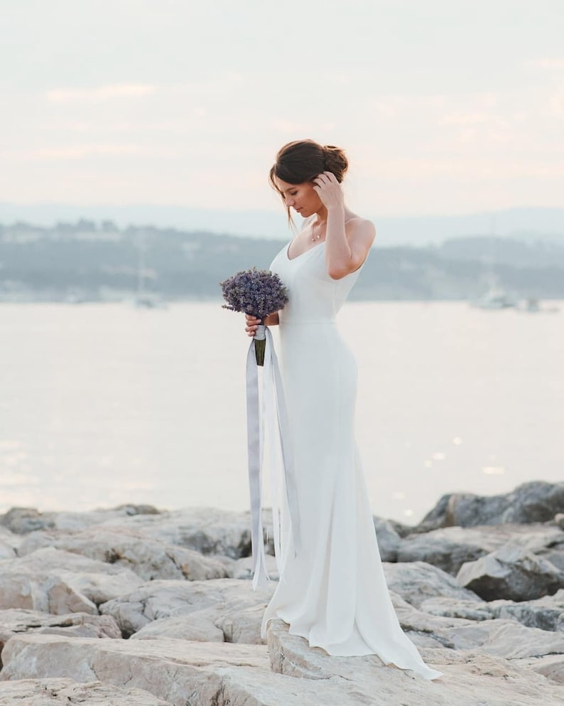 May include: A bride in a white wedding dress holds a bouquet of lavender flowers while standing on a rocky beach.