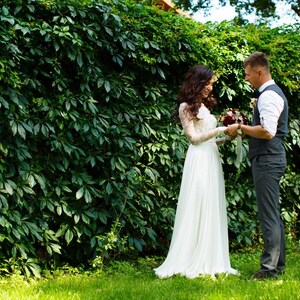 May include: A bride and groom stand in front of a green ivy wall. The bride is wearing a white wedding dress and the groom is wearing a gray suit with a vest. They are holding a bouquet of flowers.