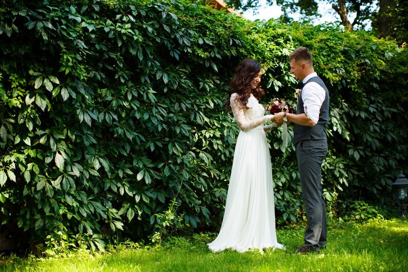 May include: A bride and groom stand in front of a green ivy wall. The bride is wearing a white wedding dress and the groom is wearing a gray suit with a vest. They are holding a bouquet of flowers.