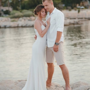 May include: A couple in white clothing standing on a rocky shore by the water. The woman is wearing a white dress and the man is wearing a white shirt and beige shorts.