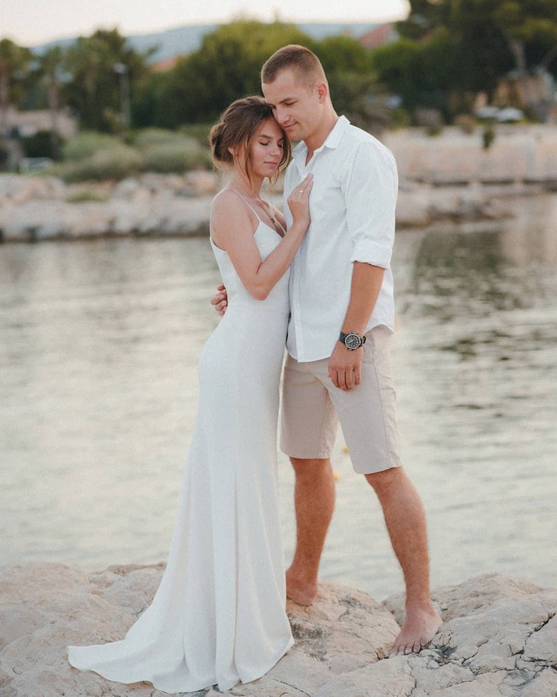 May include: A couple in white clothing standing on a rocky shore by the water. The woman is wearing a white dress and the man is wearing a white shirt and beige shorts.