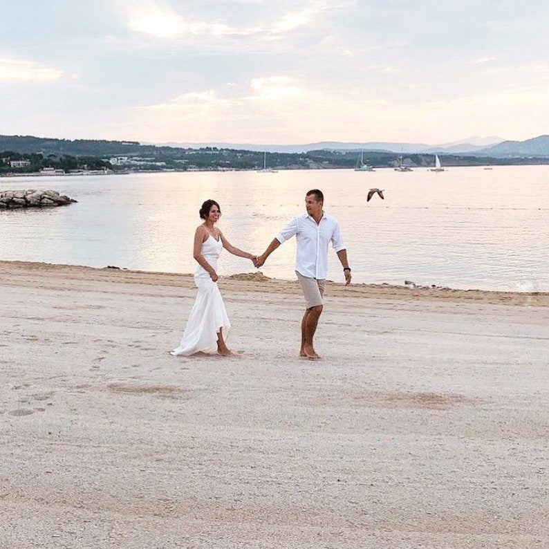 May include: A couple walks hand-in-hand on a sandy beach towards the ocean. The bride is wearing a white wedding dress and the groom is wearing a white shirt and khaki shorts. The ocean is calm and the sky is a soft blue with a few clouds.