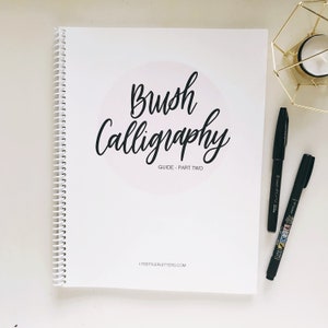 May include: A white spiral-bound workbook titled Brush Calligraphy Guide - Part Two with black text and a light pink circular accent. Two black brush pens are positioned to the right of the book, alongside a small gold geometric wire holder.