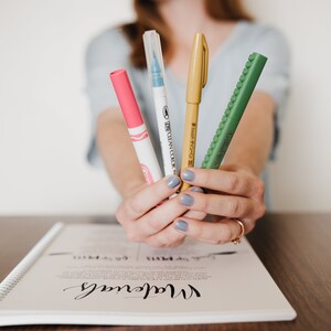 May include: Four markers held together, including a pink marker with a white cap, a clear marker with a blue tip, a gold marker with a matching cap, and a green textured marker with the text "Artline Stix" printed on the side of the barrel.