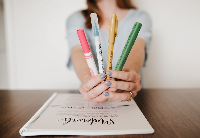 May include: Four markers held together, including a pink marker with a white cap, a clear marker with a blue tip, a gold marker with a matching cap, and a green textured marker with the text "Artline Stix" printed on the side of the barrel.