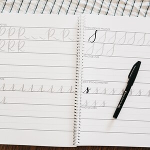 May include: A spiral-bound calligraphy practice workbook with white pages featuring black printed lines and gray letter tracing guides for the letters R and S. A black brush pen rests on the right page, which includes sections for basic strokes and practice lines.