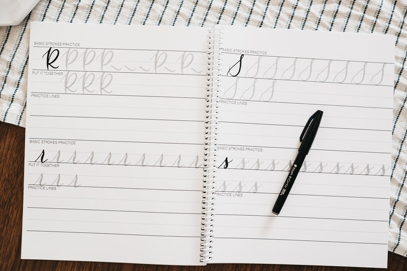 May include: A spiral-bound calligraphy practice workbook with white pages featuring black printed lines and gray letter tracing guides for the letters R and S. A black brush pen rests on the right page, which includes sections for basic strokes and practice lines.