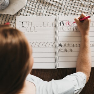 May include: A spiral-bound calligraphy practice workbook with white pages and black printed lines. The workbook features sections labeled Basic Strokes Practice and Put It Together, with handwritten letter M examples in black and red ink.