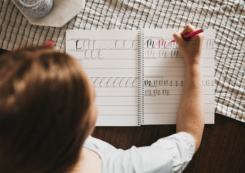 May include: A spiral-bound calligraphy practice workbook with white pages and black printed lines. The workbook features sections labeled Basic Strokes Practice and Put It Together, with handwritten letter M examples in black and red ink.