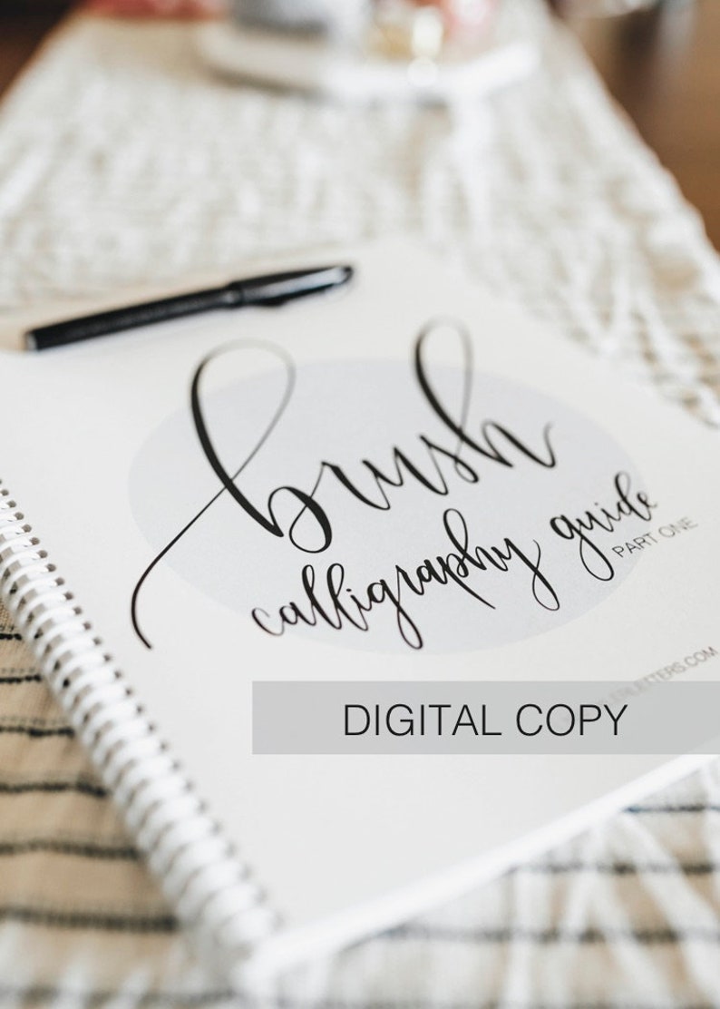 May include: A white spiral-bound brush calligraphy guide with black text that reads brush calligraphy guide part one and digital copy, accompanied by a black pen resting on the cover, featuring a light gray circular graphic element.