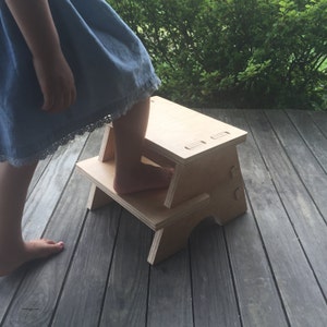 May include: A wooden two-step stool with a natural finish. A child is standing on the top step.