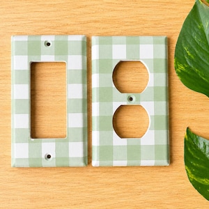 May include: Two rectangular wall plates featuring a light sage green and white gingham check pattern. One plate has a single large rectangular opening for a rocker switch, and the other has two circular openings for a standard duplex outlet.