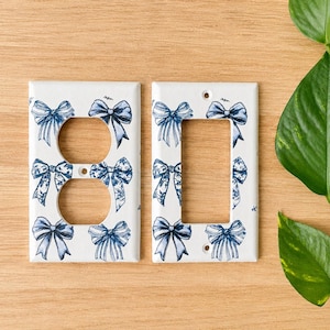 Op de afbeelding: Twee witte wandplaten voor stopcontacten met een herhalend blauw-wit strikpatroon. Eén plaat heeft twee ronde openingen voor een dubbel stopcontact, en de andere heeft een rechthoekige opening voor een wipschakelaar.