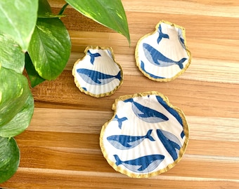 May include: Three white ceramic seashell-shaped dishes with gold trim. Each dish features a blue whale design.