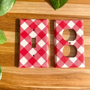 May include: Two red and white gingham patterned light switch and outlet covers. The switch cover has a single toggle switch and the outlet cover has a standard double outlet.