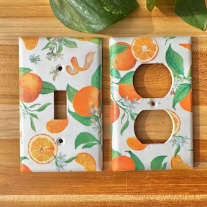 Puede incluir: Dos placas de enchufe blancas con un vibrante diseño de cítricos naranja y verde. Las placas presentan naranjas, rodajas de naranja, hojas verdes y flores blancas. Una placa tiene un interruptor, la otra dos enchufes.