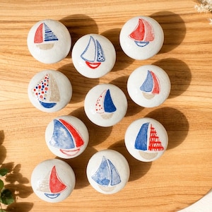Könnte beinhalten: Zehn weiße Keramik-Schrankknöpfe, jeweils handbemalt mit einem Segelboot-Design. Die Segelboote haben rote, blaue und beige Segel. Die Knöpfe sind auf einer Holzoberfläche angeordnet.