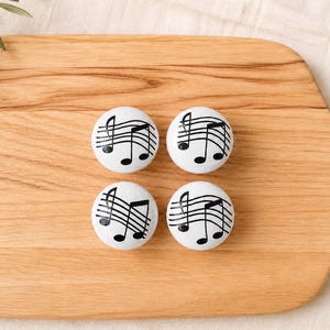 May include: A set of four round white ceramic knobs, each featuring a black printed design of a musical staff with notes. The knobs are arranged in a square formation, displaying a consistent graphic pattern across all four individual pieces.