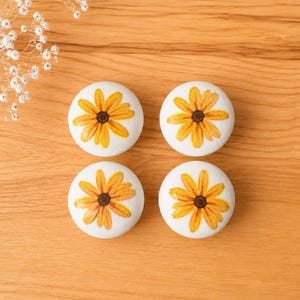 May include: Four white cabinet knobs with a yellow and brown daisy design. The knobs are arranged on a light brown wooden surface. The daisy has yellow petals and a brown center.