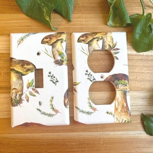 May include: Two decorative light switch covers with a mushroom design. One cover has a toggle switch opening, the other has two outlet openings. The covers are white with a detailed illustration of mushrooms and foliage.