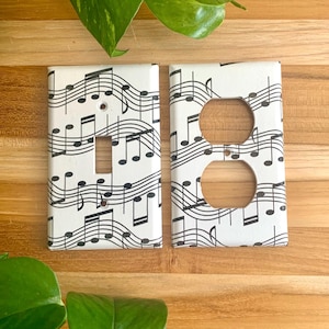Könnte beinhalten: Zwei weiße Steckdosenabdeckungen mit einem Design aus schwarzen Noten. Eine Abdeckung hat eine einzelne Schalteröffnung, die andere zwei kreisförmige Öffnungen für Steckdosen. Die Abdeckungen befinden sich auf einer Holzoberfläche.