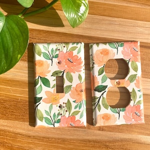 Könnte beinhalten: Zwei dekorative Lichtschalter- und Steckdosenabdeckungen mit einem Blumenmuster. Die Abdeckungen haben einen weißen Hintergrund mit rosa und orangefarbenen Blumen und grünen Blättern.
