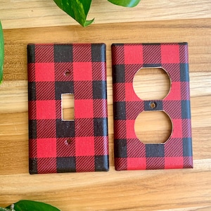 May include: Two decorative wall plates with a red and black buffalo plaid pattern. One plate has a single switch opening, and the other has two circular outlet openings. The plates are made of a durable material and are designed to cover electrical outlets and switches.