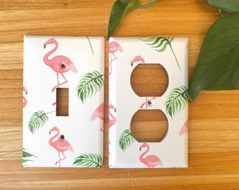 May include: Two white electrical outlet covers with a flamingo and tropical leaf pattern. One cover has a single switch opening, the other has two circular outlet openings. The design features pink flamingos and green leaves.