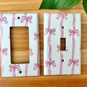May include: Two white light switch covers with a pink bow and ribbon pattern. The covers have a rectangular shape with a single switch opening and a larger opening. The background is a light wood surface.